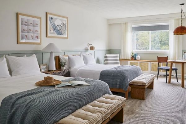 A cozy hotel room features two double beds with gray blankets, art on the walls, benches at end of the beds, and a desk area by the window.