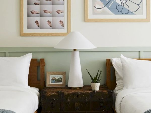 Two beds with white linens, a lamp, a small plant, and a framed photo on a shared nightstand. Two framed artworks above the heads of the beds.