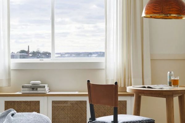 A cozy room with a window view overlooking water, featuring a table, chair, orange lamp, and books stacked on a shelf beneath the window.