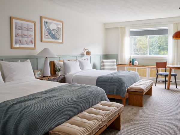 A bright hotel room with two queen beds, teal accents, a cushioned bench at each foot, a long desk by the window, and warm natural light from the window.
