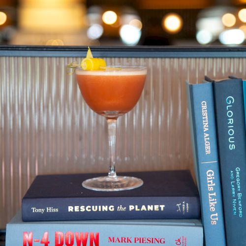 A pink cocktail with an orange twist sits in a coupe atop a stack of blue books, glow-lit background, cozy bar vibes.