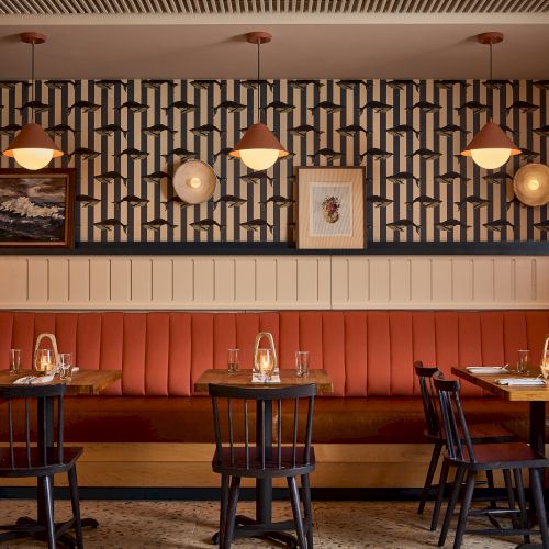 A stylish restaurant booth with red banquette seating, wooden chairs, round lamps, geometric wallpaper, and framed art above the tables.