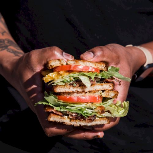 A person with tattoos holds a stacked, multi-layered burger with lettuce, tomato, cheese, and patties, ready to bite.