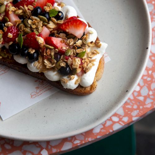 A crispy toast topped with whipped cream, fresh berries, granola, and seeds, arranged artfully on a white parchment on a pale plate.