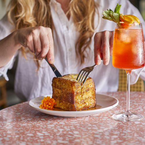 A person in a white shirt is cutting a square loaf or cake on a plate with a side of mashed potatoes or carrots, next to a bright orange cocktail garnished with mint, on a pink speckled table.