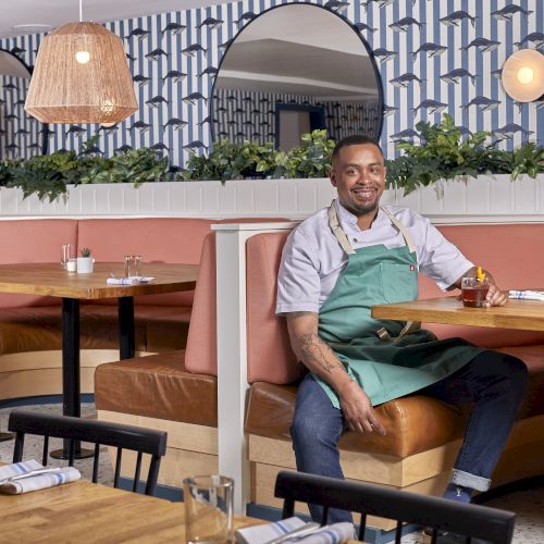 A smiling server wearing a green apron sits in a pink-booth dining area with wooden tables, plants, and warm lighting, ready to serve.