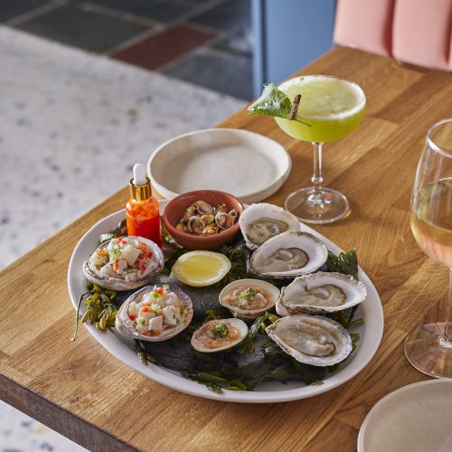 A delectable plate of oysters and tapas with sauces, lemon, and garnishes, plus a white wine cocktail and a glass of white wine on a wooden table.