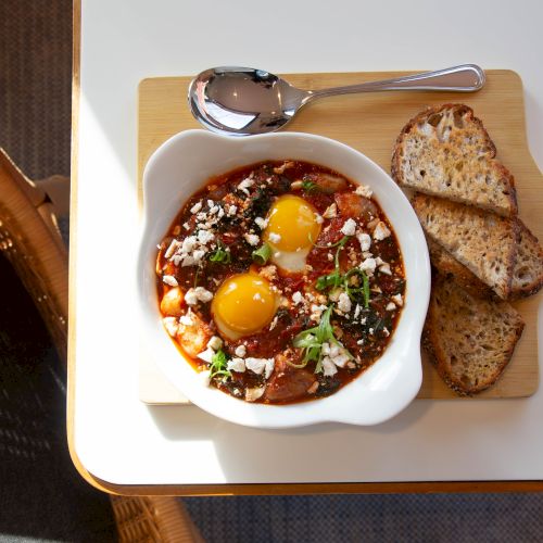 A white bowl of chili/tajarin topped with two eggs, herbs, cheese, and corn, served with several slices of crusty bread on a wooden board.