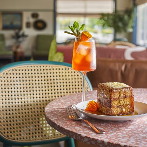 A glass of orange soda with a mint garnish beside a plated cake on a pink terrazzo table in a cozy cafe.