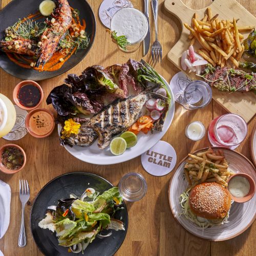 A spread of grilled steak, burgers, fries, salads, tacos, and drinks on a wooden table&mdash;colorful, hearty dishes for a big group.