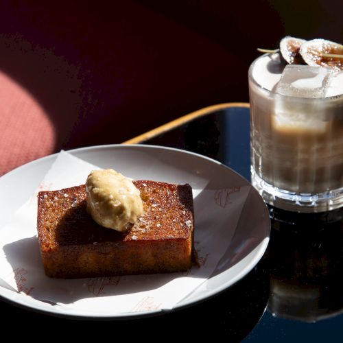 A slice of cake on a white plate with a dollop of cream, and a frothy iced coffee with marshmallows on a dark glass table.