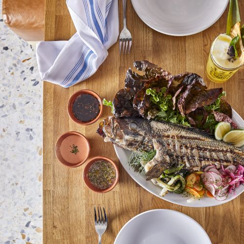 Grilled whole fish with herbs on a plate, lemon wedges, sides of veggies, sauces, and beverages on a wooden table set for a meal.