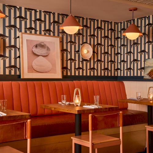 A chic dining area with warm orange booth seating, wooden tables, geometric-patterned wallpaper, pendant lights, and framed art. end at 140 chars.