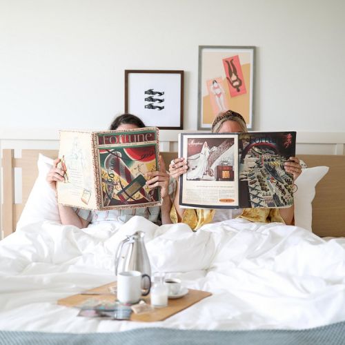 A cozy bedroom scene with a bed, pillows, and a person lounging among soft blankets, framed art on the wall behind, and warm natural light.
