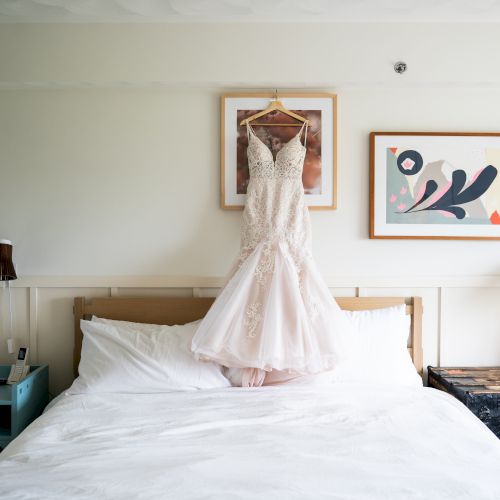 A sunlit hotel room with a white bedspread, a girl in a pink dress posing in the center, and a serene blue-green wall behind.