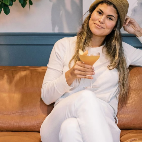 A woman wearing white sits on a brown couch, holding an orange drink and smiling, with a blue wall in the background.