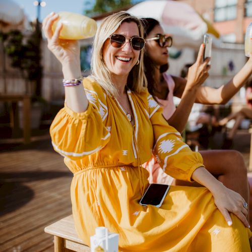 A woman in a yellow dress sits outdoors with sunglasses, smiling and holding a drink, sunny terrace backdrop.