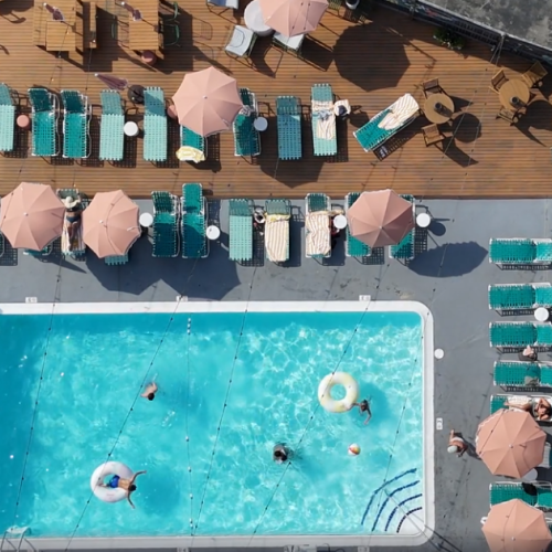 Aerial view of a resort pool area with a rectangular blue pool, sun loungers lined up, pink umbrellas, and guests swimming or lounging both in and around the pool.