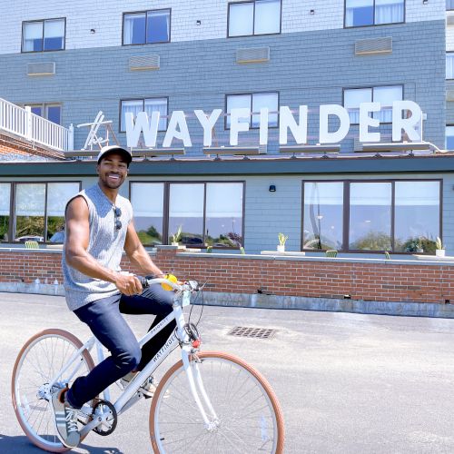 A person rides a bicycle on a street, passing by a building with a sign that reads &ldquo;WAYFINDER,&rdquo; under a clear sky.