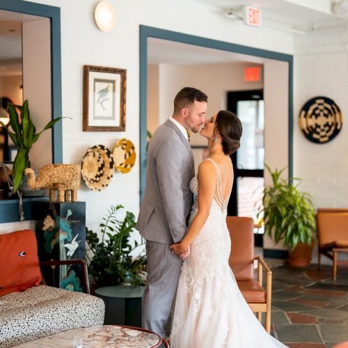 A bride in a white wedding gown stands in a cozy, well‑lit living room with plants, artwork, and family photos.
