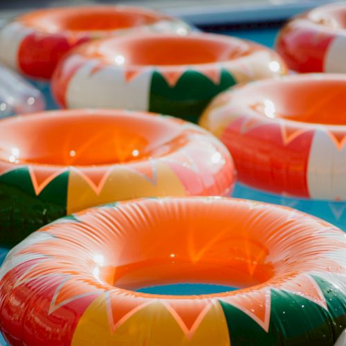 Colorful inflatable pool toys/inner tubes floating together, bright oranges, greens, and pinks, outdoors in a sunny scene.