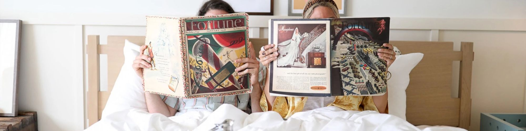 Two people are lying in bed, holding up large magazines that cover their faces.