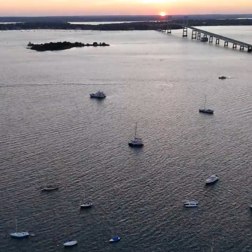 A calm harbor at sunset with numerous small boats anchored, a long pier extending toward the horizon, and a tiny rocky island nearby, serene.