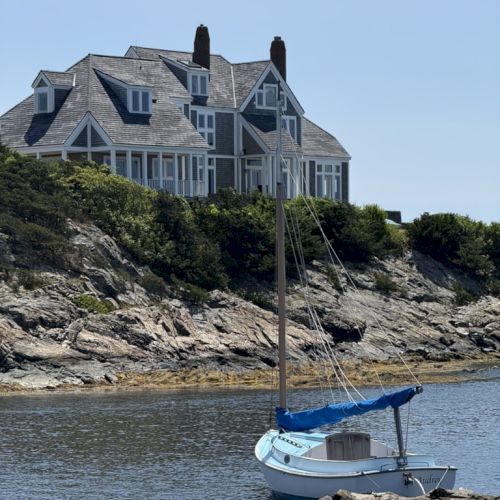 A grand coastal house perched on rocky shore, with chimneys and multiple gables, overlooking a calm bay and a small sailboat grounded on the rocky beach.