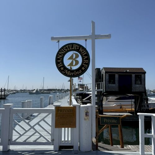 A sunny marina entrance with a white gate, signboard, and a small wooden building beside calm boats and clear blue water.
