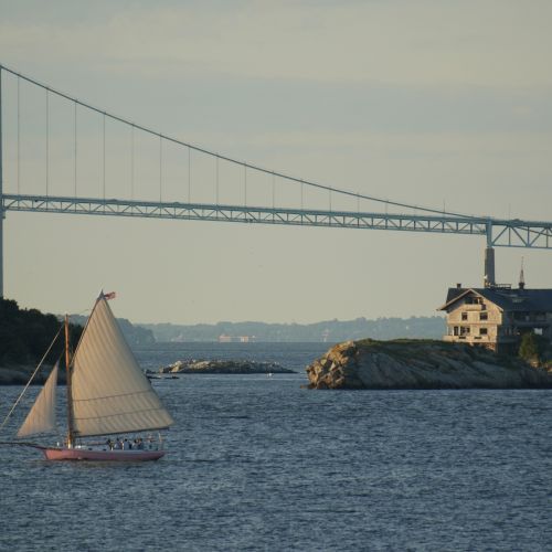 A sailboat glides past a small island with a historic house, under a long suspension bridge spanning the water.