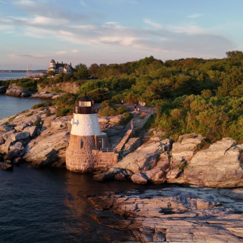 A rocky coastline with a small lighthouse perched on a stone jetty, surrounded by greenery and calm blue water at sunset.