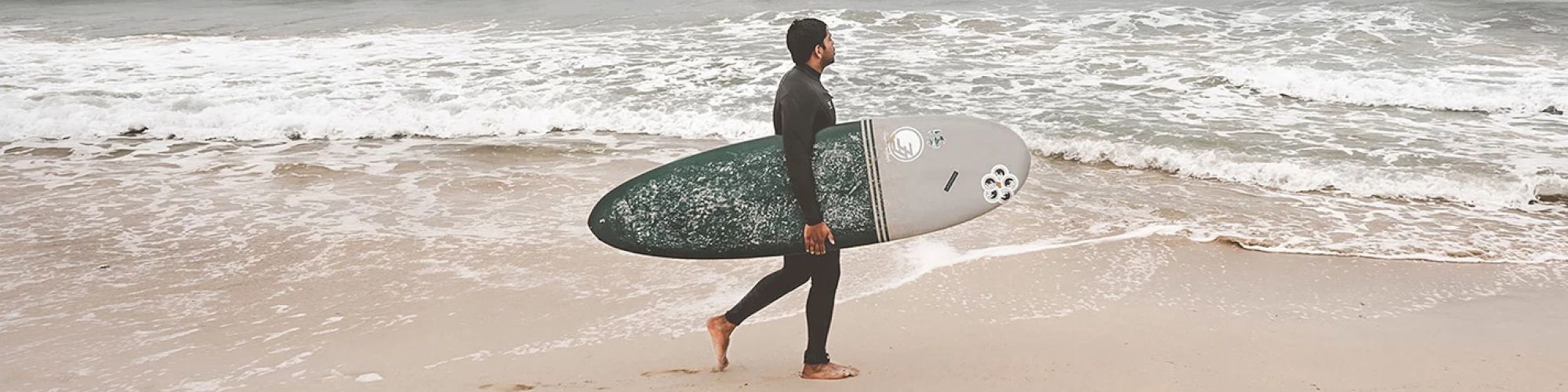 A person wearing a wetsuit walks barefoot on the beach while carrying a surfboard, with waves and the ocean in the background.