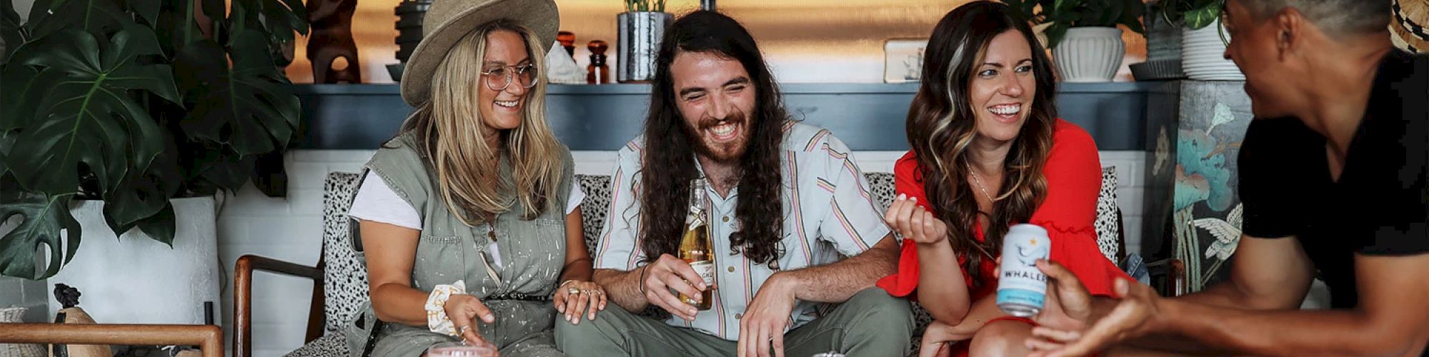 Four people are sitting on a couch, laughing and enjoying drinks in a cozy, plant-decorated space.