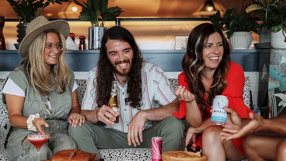 Four people are sitting on a couch, laughing and enjoying drinks in a cozy, plant-decorated space.