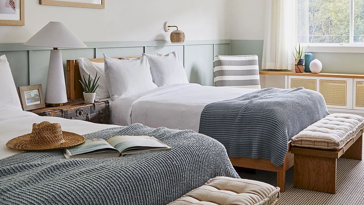 The image shows a cozy hotel room with two beds, a wooden table with chairs, a bench, a hat, an open book, plants, and framed pictures on the wall.