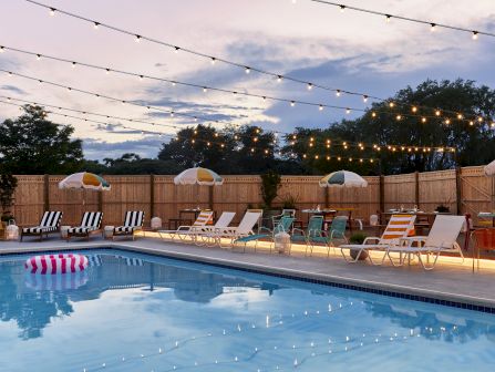 A nighttime pool area with string lights, lounge chairs, umbrellas, and a calm blue pool reflecting the lights.