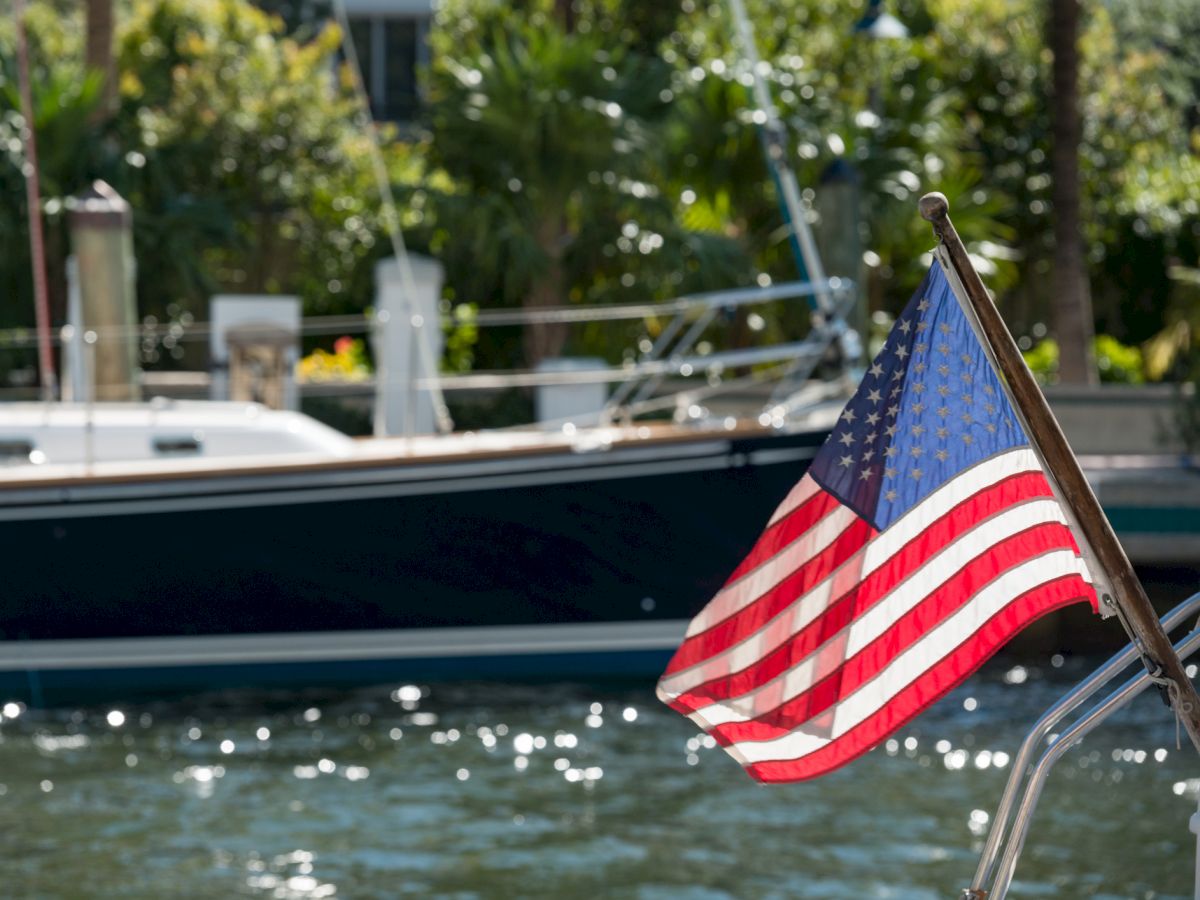 A U.S. flag is seen waving on a boat, with another boat and greenery in the background, reflecting a peaceful water scene.