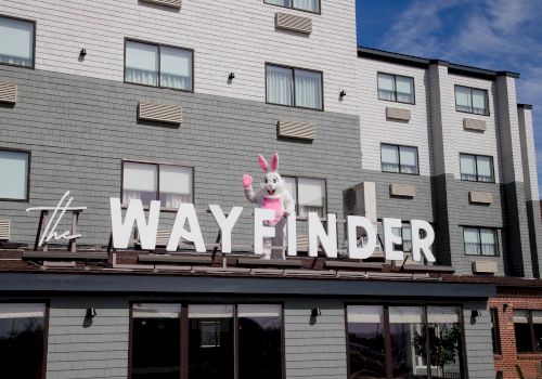 A storefront sign that reads &ldquo;The Wayfinder&rdquo; with a pink bunny statue on top of the letters, in front of a gray building.