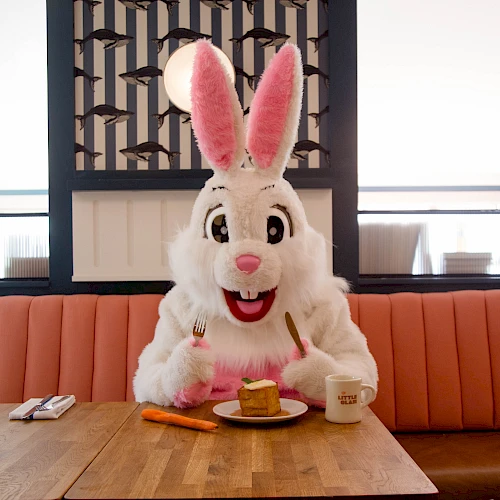 A person in a bunny costume sits at a table with a small pancake lunch, pink napkins, and a mug, in a cozy restaurant.