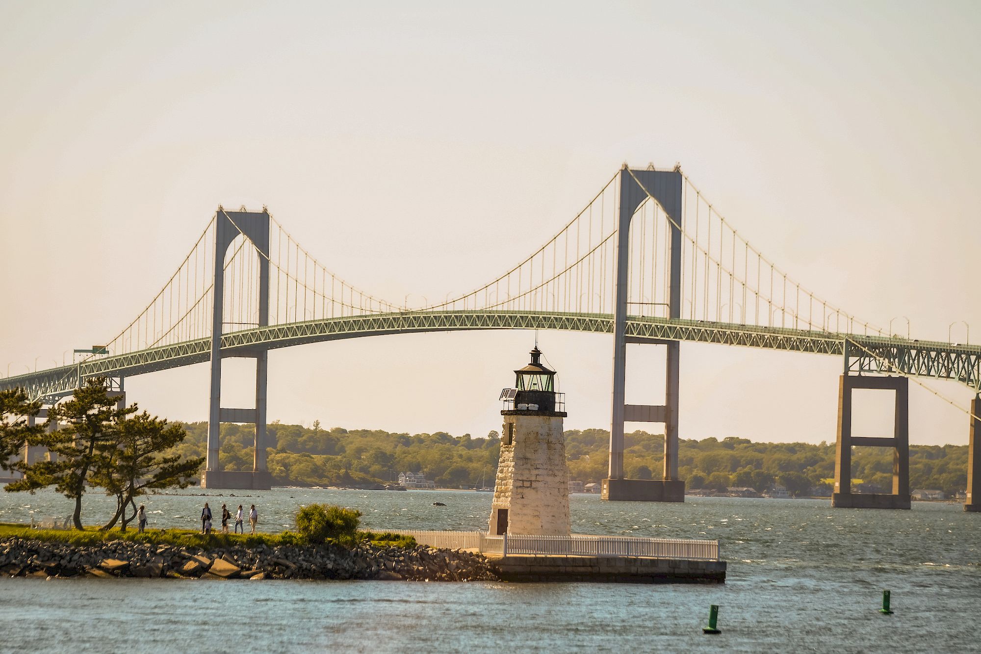 A bridge spans a harbor with a small lighthouse on a rocky jetty, calm water, and distant trees under a pale sky.