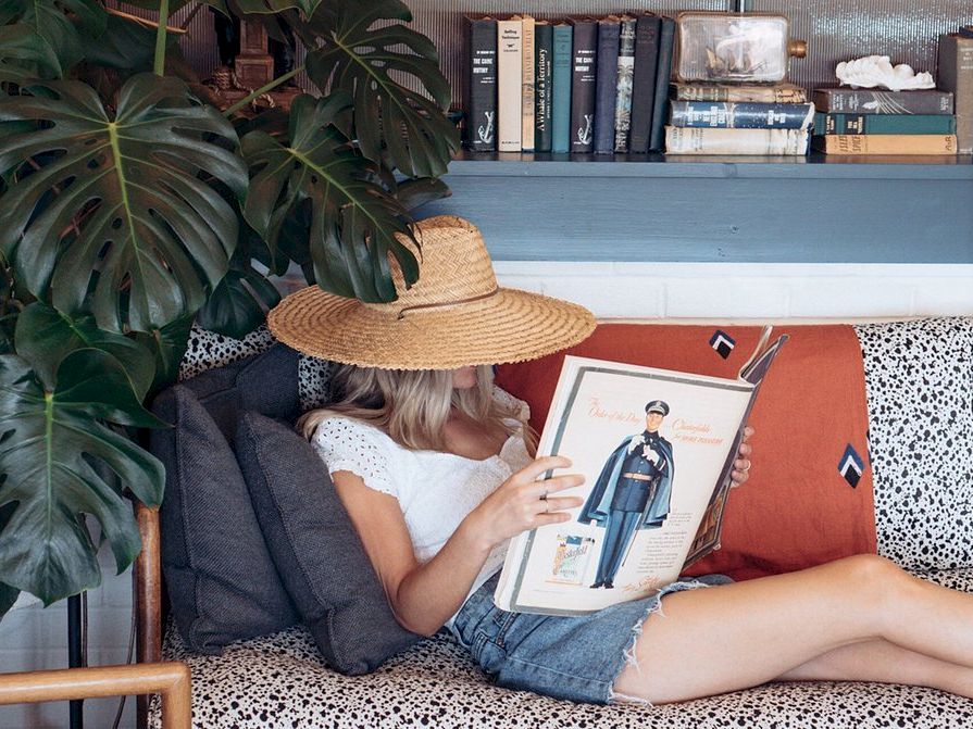 A person wearing a straw hat lounges on a patterned couch, reading a fashion illustration book, with plants and bookshelves in the background.