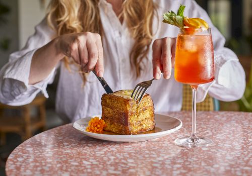 A person at a table cutting into a square pastry with a side of carrot puree, plus a bright orange cocktail garnished with mint.