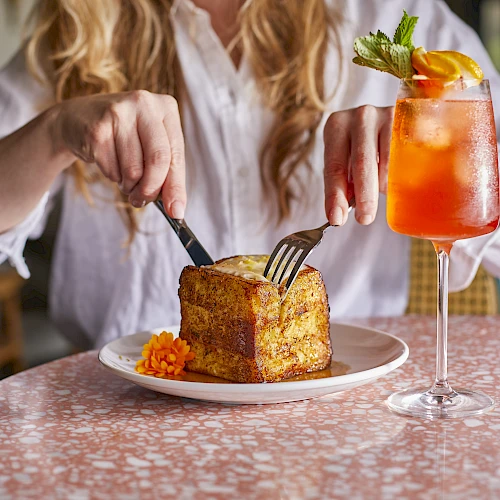 A person at a table cutting into a square pastry with a side of carrot puree, plus a bright orange cocktail garnished with mint.