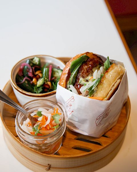 A bao-style sandwich with greens, served on a round wooden tray alongside a small salad and pickled veggies, all on a white table.