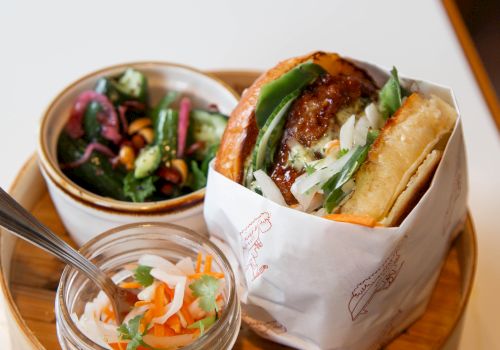 A bao-style sandwich with greens, served on a round wooden tray alongside a small salad and pickled veggies, all on a white table.