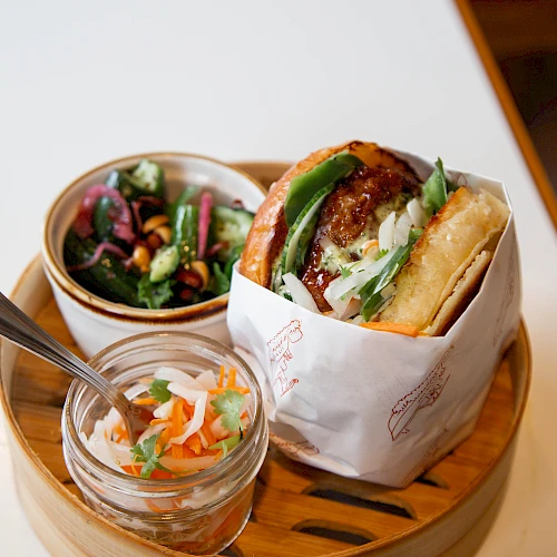 A bao-style sandwich with greens, served on a round wooden tray alongside a small salad and pickled veggies, all on a white table.