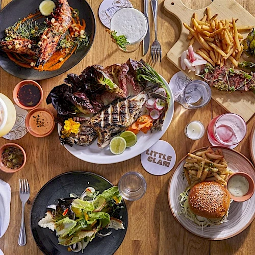 A top-down view of a colorful restaurant table with various dishes: burgers, fries, grilled meats, salads, sauces, and drinks arranged on plates and boards, inviting shared dining.