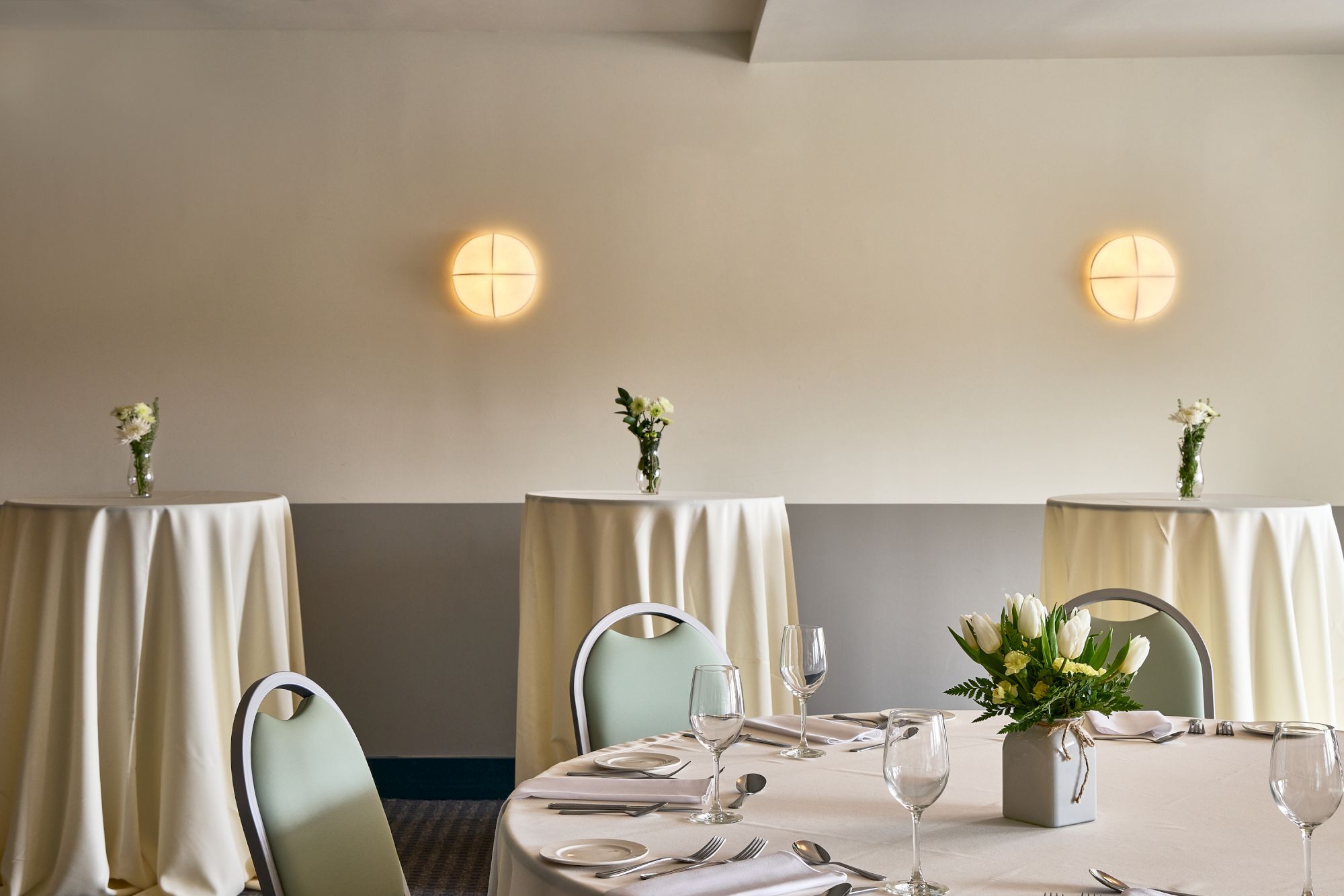 A set banquet table with place settings, fresh white flowers in vases, and two warm round wall lights in a meeting hall.