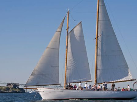 A sailboat with white sails and several people on board, sailing near a small island and a bridge in the background, with a flag at the stern.