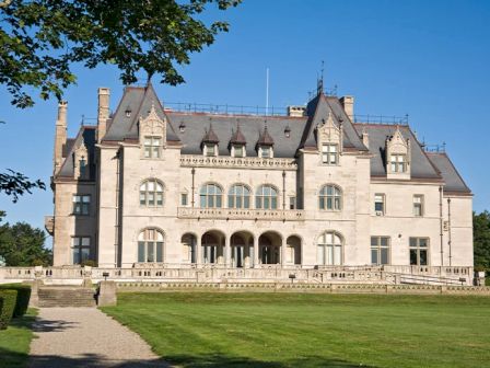 A large, ornate mansion with multiple windows and peaked roofs, surrounded by a well-maintained lawn under a clear blue sky.